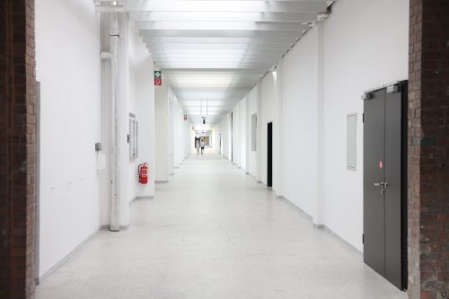Langer, heller Flur mit weißen Wänden, einigen Türen und einem Feuerlöscher an der Wand; am Ende sind Menschen zu sehen.