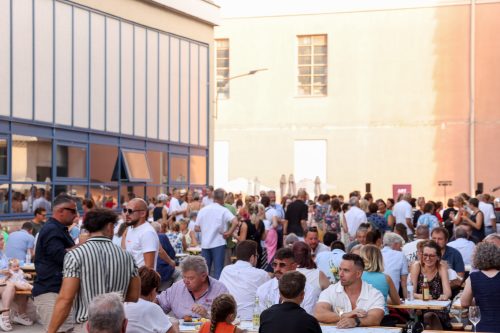 Viele Menschen sitzen und stehen auf einem sonnigen Innenhof bei einem geselligen Fest mit Speisen und Getränken.