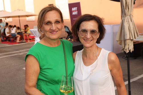 Zwei lächelnde Frauen, eine in grünem Kleid und eine in weißer Bluse, stehen mit einem Glas Wein auf einer Außenterrasse.