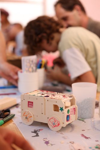 Selbstgebasteltes Fahrzeug aus Holz mit bunten Verzierungen auf einem Tisch, im Hintergrund Kinder beim Basteln.