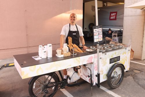 Ein Mann steht lächelnd hinter einem dekorierten Eiswagen mit Eiswaffeln, Bechern und dem Schild „LEONES GELATO“.