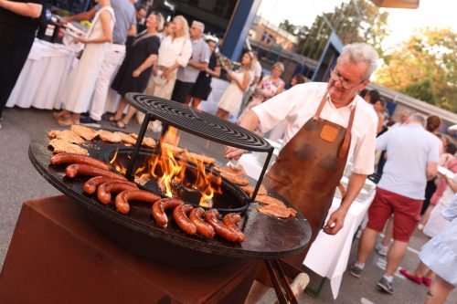 Ein Mann grillt Würste und Fleischscheiben auf einer großen Feuerplatte bei einer Veranstaltung mit mehreren Gästen im Freien.