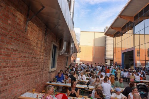 Viele Menschen sitzen an langen Tischen im Freien und genießen gemeinsam Essen und Trinken bei sonnigem Wetter.