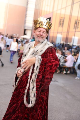 Ein lächelnder Mann trägt eine königliche Robe aus rotem Samt mit Pelzbesatz und eine goldene Krone, während er im Freien steht.