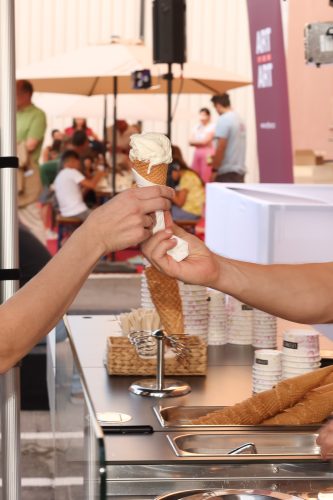 Zwei Personen übergeben sich eine große Waffel mit Vanilleeis an einem Eisstand im Freien.