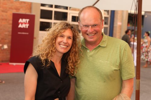 Eine Frau mit lockigem Haar und ein Mann mit Brille lächeln gemeinsam in die Kamera auf einer Veranstaltung.