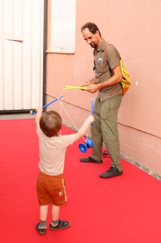 Ein Kind spielt mit einem Diabolo, während ein erwachsener Mann mit gelbem Rucksack zuschaut und hilft.