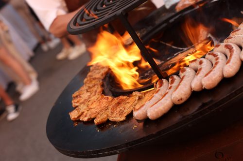 Würste und Fleischstücke werden auf einem runden Grill über offener Flamme im Freien gegrillt.