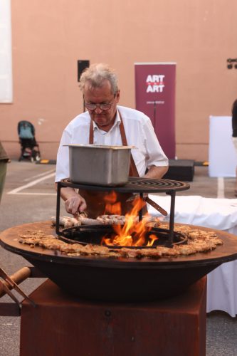 Ein älterer Mann grillt Fleisch auf einer offenen Feuerstelle im Freien, im Hintergrund ist ein "ART ART"-Schild zu sehen.