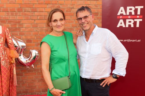 Eine Frau im grünen Kleid und ein Mann im weißen Hemd lächeln vor einer Wand mit dem Schriftzug „ART FOR ART“.