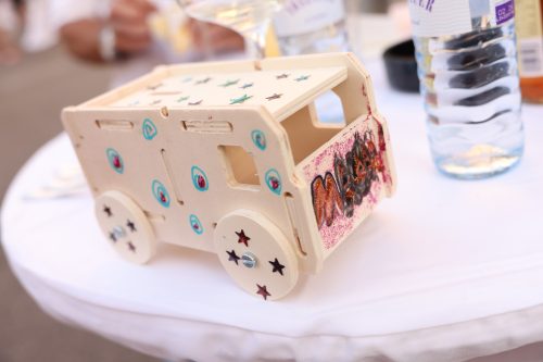 Holzspielzeugauto, verziert mit bunten Aufklebern und dem Namen "Melek" an der Seite, steht auf einem weißen Tisch neben einer Wasserflasche.