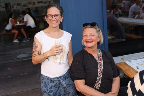 Zwei Frauen lächeln in die Kamera, eine steht mit einem Getränk in der Hand, die andere sitzt an einem Tisch draußen.