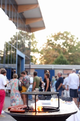 Grill mit Würstchen im Vordergrund, im Hintergrund eine Gruppe von Menschen bei einer Veranstaltung im Freien.