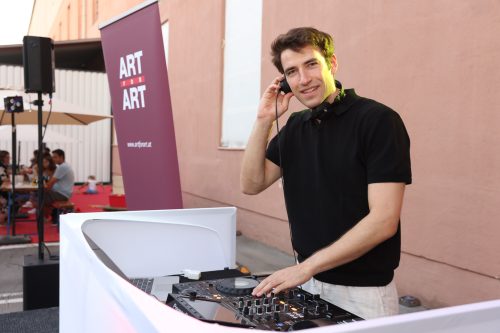 Ein DJ steht an einem Mischpult im Freien und lächelt, während er Musik auf einer Veranstaltung mit Kunstbanner im Hintergrund auflegt.