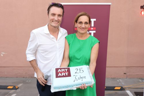 Ein Mann und eine Frau halten gemeinsam eine Torte mit der Aufschrift "25 Jahre ART" vor einem ART-Logo-Banner.