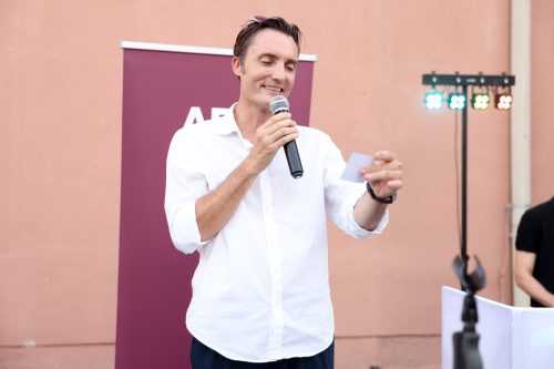 Ein Mann in weißem Hemd spricht mit einem Mikrofon und hält einen Zettel, im Hintergrund ein weinrotes Banner.