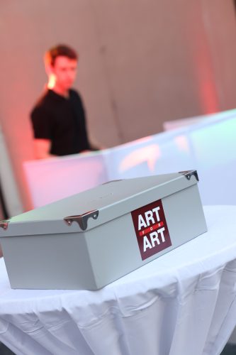 Graue Metallbox mit Aufkleber "ART FOR ART" auf einem runden, weiß gedeckten Tisch, unscharfe Person im Hintergrund.