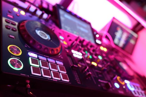 Ein DJ-Mischpult mit leuchtenden Tasten und Reglern vor unscharfen Monitoren in einer pink beleuchteten Umgebung.