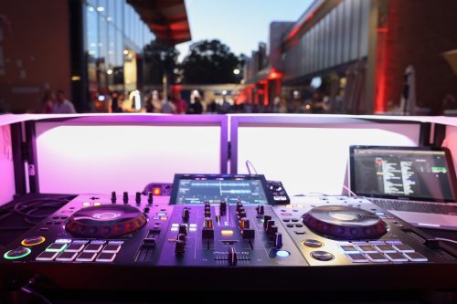 DJ-Set mit Mischpult, Laptop und bunten Lichtern im Freien, im Hintergrund Menschen auf einer Veranstaltung bei Abenddämmerung.
