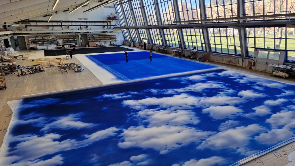In einer großen Halle liegen bemalte Bühnenhintergründe mit Wolkenmotiven auf dem Boden, zwei Personen arbeiten daran.