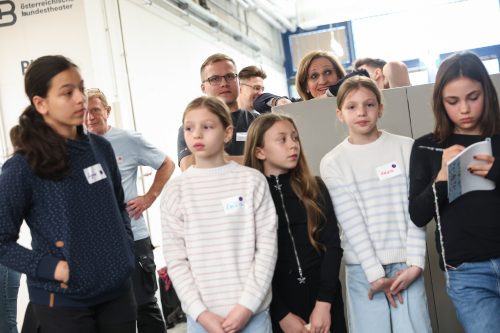 Eine Gruppe von Kindern und Erwachsenen steht zusammen in einem hellen Raum; einige Kinder tragen Namensschilder.