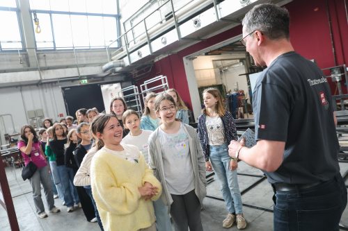 Eine Gruppe von Kindern steht in einer Werkhalle und hört aufmerksam einem Erwachsenen zu, der ihnen etwas erklärt.