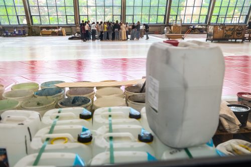 Eine Gruppe von Menschen steht in einer Halle zusammen, im Vordergrund sind viele Kanister und Farbdosen zu sehen.