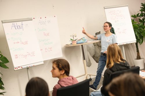 Eine Frau hält einen Vortrag vor einer kleinen Gruppe; neben ihr stehen Flipcharts mit handgeschriebenen Notizen zum Tagesablauf.