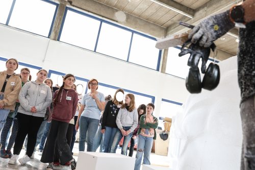 Eine Gruppe von Schülerinnen beobachtet aufmerksam eine Person mit Schutzhandschuhen und Werkzeugen in einem hellen Raum.