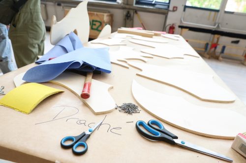 Werkbank mit ausgeschnittenen Holzformen, Schere, Nägel, Stoffresten und dem handschriftlichen Wort „Drache“ auf Papier.