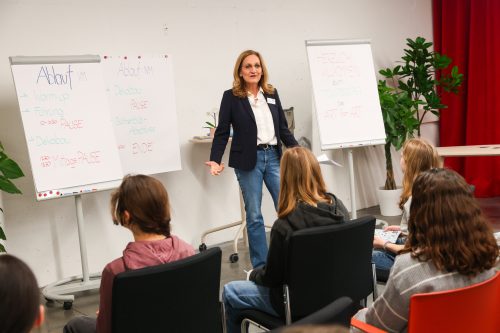 Eine Frau steht vor einer Gruppe von Jugendlichen und präsentiert, neben ihr zwei Whiteboards mit Notizen und Begrüßungstext.