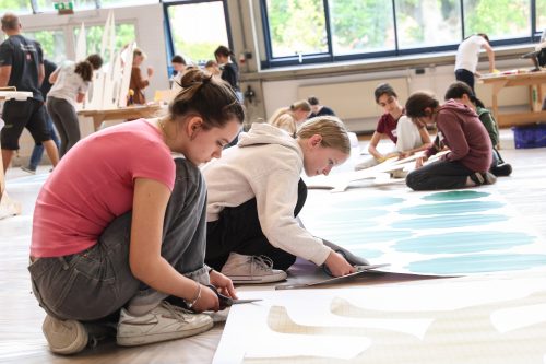 Mehrere Jugendliche sitzen am Boden und schneiden Papierformen für ein gemeinsames Kunstprojekt in einem hellen Raum aus.