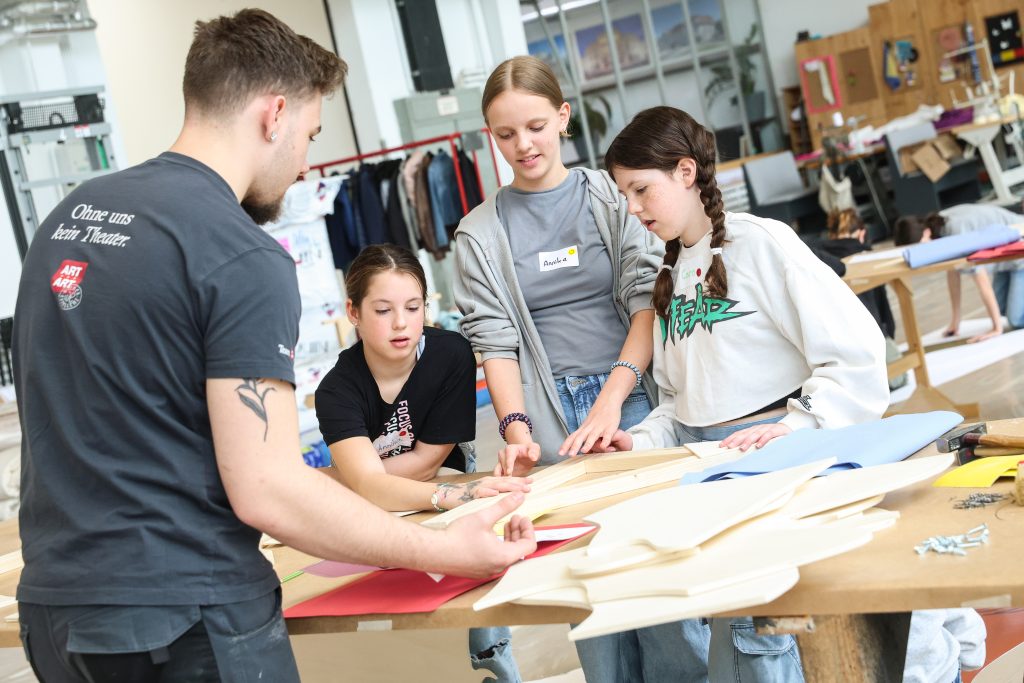 Vier Jugendliche stehen um einen Tisch und arbeiten gemeinsam an Bastelprojekten mit Papier und Holz in einem Werkraum.
