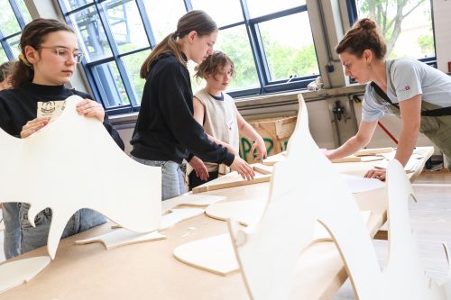Drei Jugendliche und eine Erwachsene arbeiten gemeinsam an großen, ausgeschnittenen Holzplatten in einem hellen Werkraum.