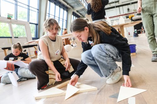 Zwei Mädchen arbeiten gemeinsam an einem Bastelprojekt auf dem Boden, während weitere Kinder im Hintergrund kreativ beschäftigt sind.