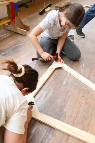 Zwei Kinder arbeiten gemeinsam mit Hammer und Nägeln an einer hölzernen Konstruktion auf einem Holzboden.