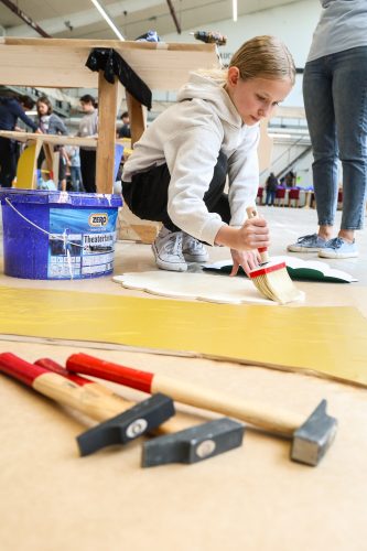 Ein Kind streicht mit einem Pinsel weiße Farbe auf eine Holzplatte; im Vordergrund liegen Hämmer und im Hintergrund arbeiten weitere Personen.