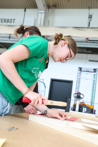 Ein Mädchen mit Brille hämmert mit einem Hammer einen Nagel in ein Stück Holz, während eine andere Person danebensteht.