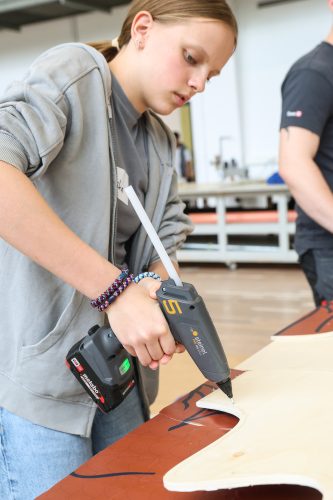 Ein Mädchen benutzt eine Heißklebepistole, um ein Stück Holz an einem Werkstatt-Tisch zu bearbeiten.