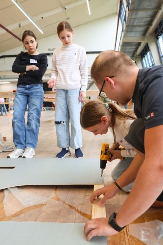 Zwei Mädchen beobachten, wie ein Mädchen zusammen mit einem Mann mit einer Bohrmaschine an einem Holzstück arbeitet.
