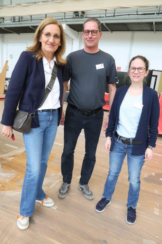 Drei Menschen stehen lächelnd nebeneinander in lässiger Kleidung in einer hellen Halle.