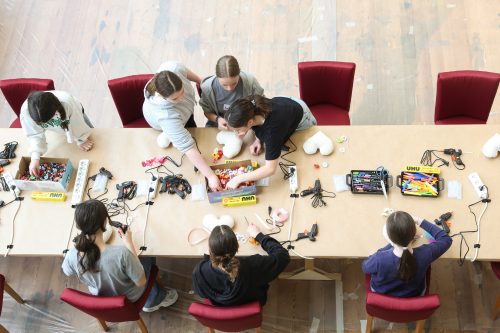 Sieben Kinder sitzen an einem Tisch mit Bastelmaterialien, Klebepistolen, Stoffherzen und verschiedenen Bastelutensilien.