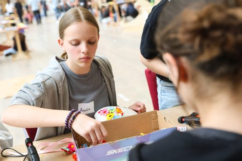 Ein Mädchen bastelt konzentriert mit bunten Materialien an einem Tisch, während weitere Personen im Hintergrund beschäftigt sind.
