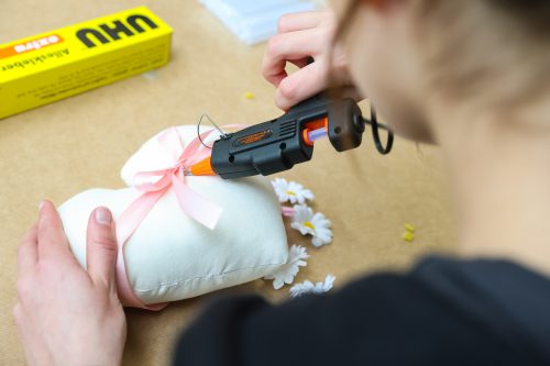 Eine Person klebt mit einer Heißklebepistole eine rosafarbene Schleife auf ein herzförmiges Kissen, daneben liegen künstliche Blumen.