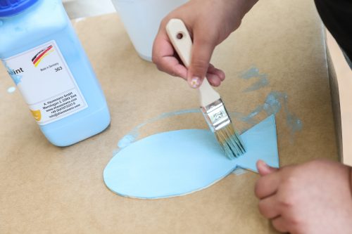 Ein Kind bemalt eine ausgeschnittene Papierform mit blauer Farbe und einem Pinsel.