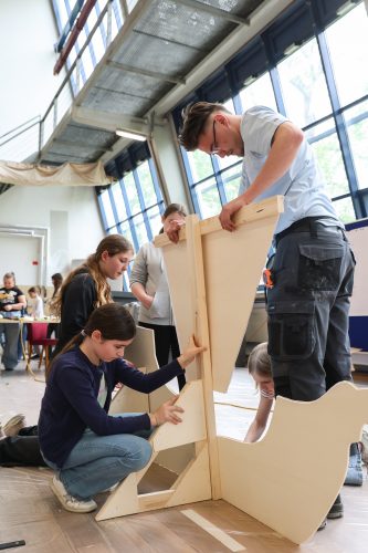 Mehrere Jugendliche und ein Erwachsener bauen gemeinsam ein großes Holzstück in einem hellen Raum mit großen Fenstern zusammen.