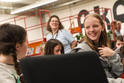 Mehrere Kinder sitzen zusammen und unterhalten sich lachend, im Hintergrund steht eine erwachsene Frau und beobachtet die Szene lächelnd.
