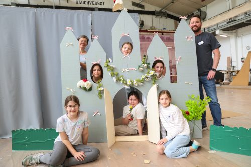 Sieben Menschen, darunter Kinder und Erwachsene, posieren fröhlich hinter und vor einer Kulisse in Form eines dekorierten Schlosses.