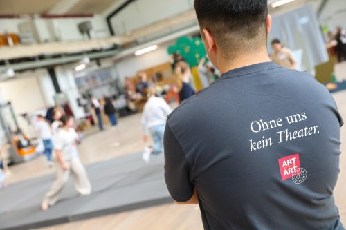 Mann mit T-Shirt-Aufdruck „Ohne uns kein Theater.“ steht in einer Probenhalle, während Menschen im Hintergrund üben.
