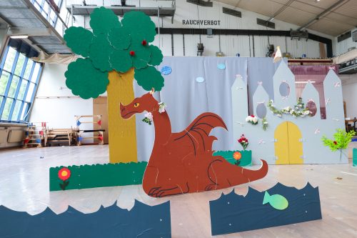Große Pappfigur eines roten Drachen vor einer Burg, neben einem Baum und einer Wiese, mit Dekorationen wie Blumen und Fischen.
